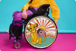 With a tuquoise wall and purple floor a Woman wears yellow top and purple trousers in a wheelchair showing the Lion King design